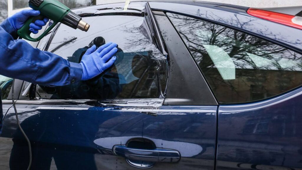 A person using a heat gun to tint a car window, applying pressure with their gloved hand.